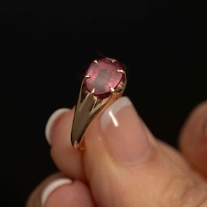 May include: A gold ring featuring a single oval-cut deep red gemstone set in a six-prong mounting. The band is smooth and polished, tapering slightly toward the setting. The gemstone displays a vibrant crimson hue with visible internal facets.