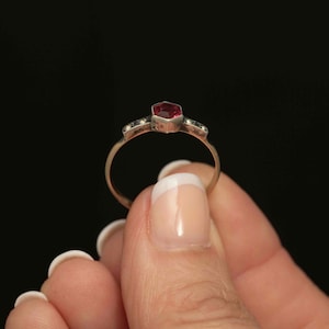 May include: A gold ring featuring a hexagonal, deep red gemstone as the centerpiece. The ring has small, decorative stones on either side of the main stone. The ring is held against a black background.