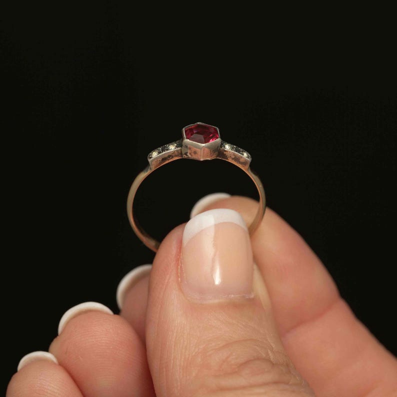May include: A gold ring featuring a hexagonal, deep red gemstone as the centerpiece. The ring has small, decorative stones on either side of the main stone. The ring is held against a black background.