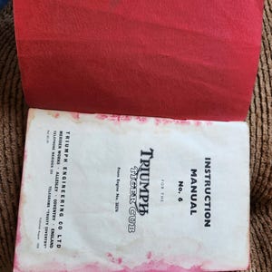 May include: An open vintage instruction manual for a Triumph Tiger Cub motorcycle. The cover is white with black text and the title "TRIUMPH TIGER CUB". The manual is open to reveal a red cover. The manual is slightly stained.