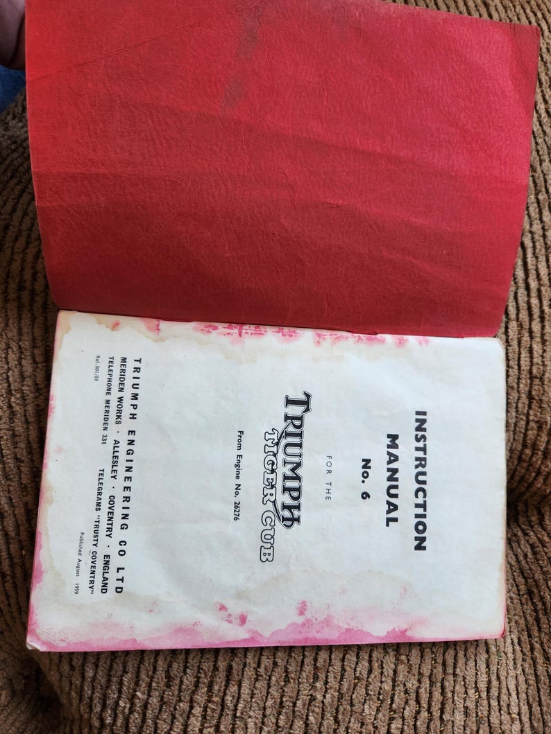 May include: An open vintage instruction manual for a Triumph Tiger Cub motorcycle. The cover is white with black text and the title "TRIUMPH TIGER CUB". The manual is open to reveal a red cover. The manual is slightly stained.