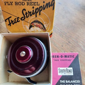 May include: A vintage OREN-O-MATIC fly rod reel in its original box. The reel is a deep burgundy color with a silver handle. The box is yellow with red and black text, and features a fish illustration. A pink and black pamphlet is also included.
