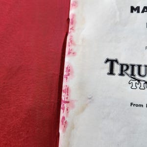 May include: A close-up of a vintage instruction manual with the word "TRIUMPH" in a stylized font. The manual is on a white background with red accents and text. The left side of the image is a solid red.