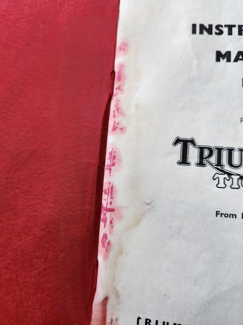 May include: A close-up of a vintage instruction manual with the word "TRIUMPH" in a stylized font. The manual is on a white background with red accents and text. The left side of the image is a solid red.