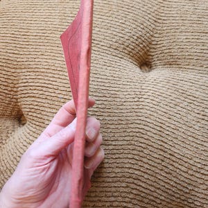 May include: A pink, rectangular book is held in a person's hand. The book's pages are visible, and the cover appears to be a solid pink color. The background is a textured, tan-colored surface.