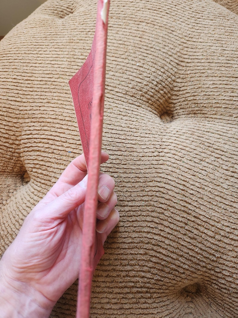 May include: A pink, rectangular book is held in a person's hand. The book's pages are visible, and the cover appears to be a solid pink color. The background is a textured, tan-colored surface.