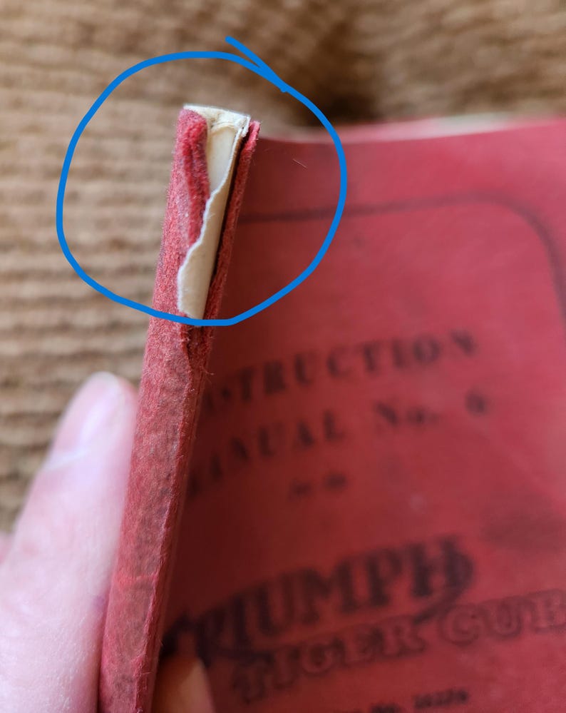 May include: Close-up of a vintage red instruction manual. The cover is worn, with visible wear and tear at the edges. The text "TRIUMPH TIGER CUB" is partially visible. The manual appears to be old and well-used.