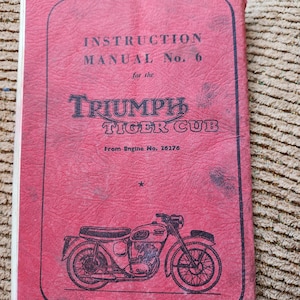 May include: A vintage instruction manual for a Triumph Tiger Cub motorcycle. The cover is a faded red with black text and a line drawing of the motorcycle. The text reads "Instruction Manual No. 6" and "Triumph Tiger Cub."