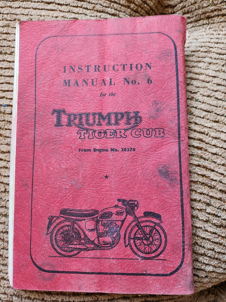 May include: A vintage instruction manual for a Triumph Tiger Cub motorcycle. The cover is a faded red with black text and a line drawing of the motorcycle. The text reads "Instruction Manual No. 6" and "Triumph Tiger Cub."
