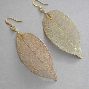 May include: A pair of gold-colored leaf earrings. The earrings are shaped like large leaves with intricate vein detailing. The earrings have a hook closure and are made of a lightweight material.