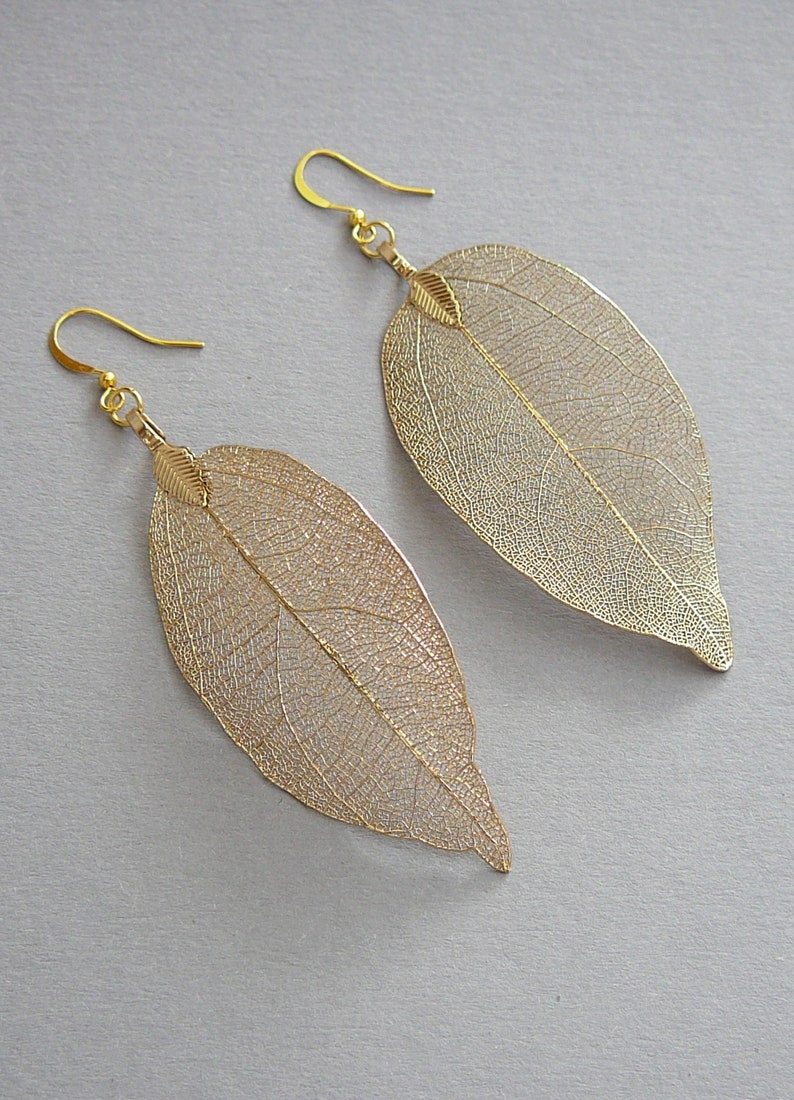 May include: A pair of gold-colored leaf earrings. The earrings are shaped like large leaves with intricate vein detailing. The earrings have a hook closure and are made of a lightweight material.