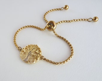 May include: A gold adjustable bracelet with a round charm featuring a detailed design of a tree with small clear stones. The bracelet has a sliding clasp.
