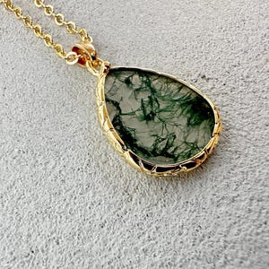 May include: A teardrop-shaped moss agate pendant set in a gold-toned metal frame with a textured edge, suspended from a matching gold-toned chain. The stone features translucent grey tones with distinct dark green, fern-like dendritic inclusions.
