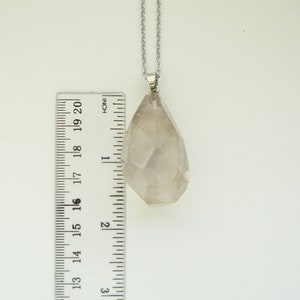 Op de afbeelding: Een heldere kwartskristal hanger aan een zilveren ketting. De hanger is ongeveer 5 cm lang en heeft een gefacetteerde vorm. De kristal is helder en heeft een lichte glans.