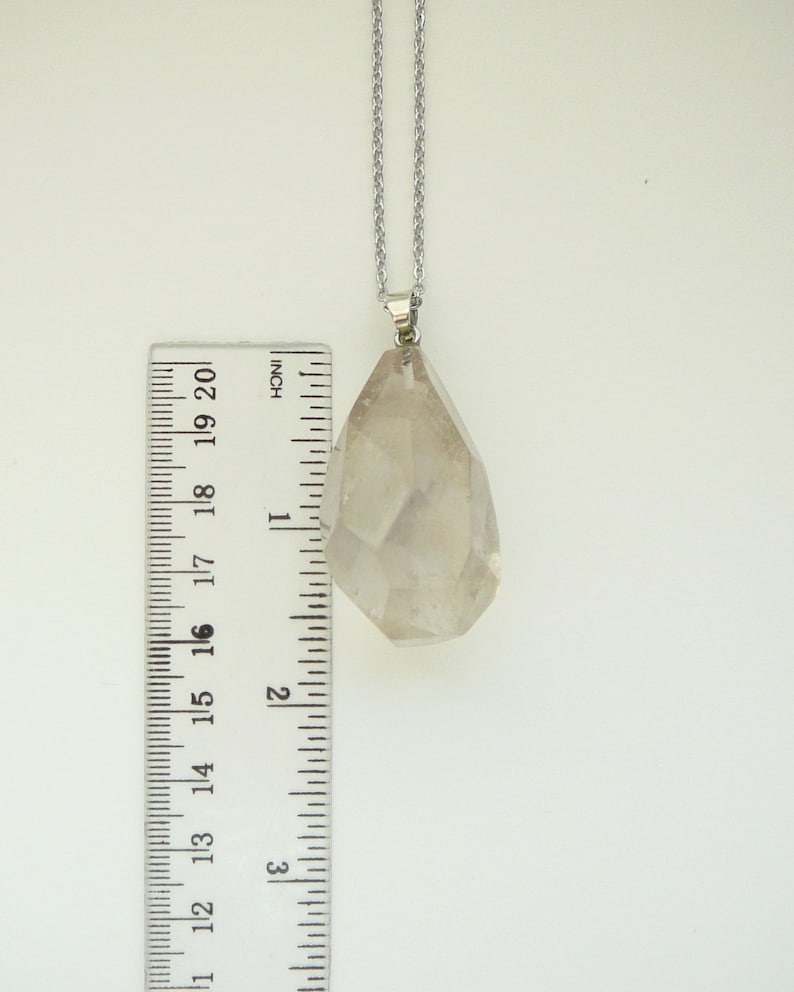 Op de afbeelding: Een heldere kwartskristal hanger aan een zilveren ketting. De hanger is ongeveer 5 cm lang en heeft een gefacetteerde vorm. De kristal is helder en heeft een lichte glans.