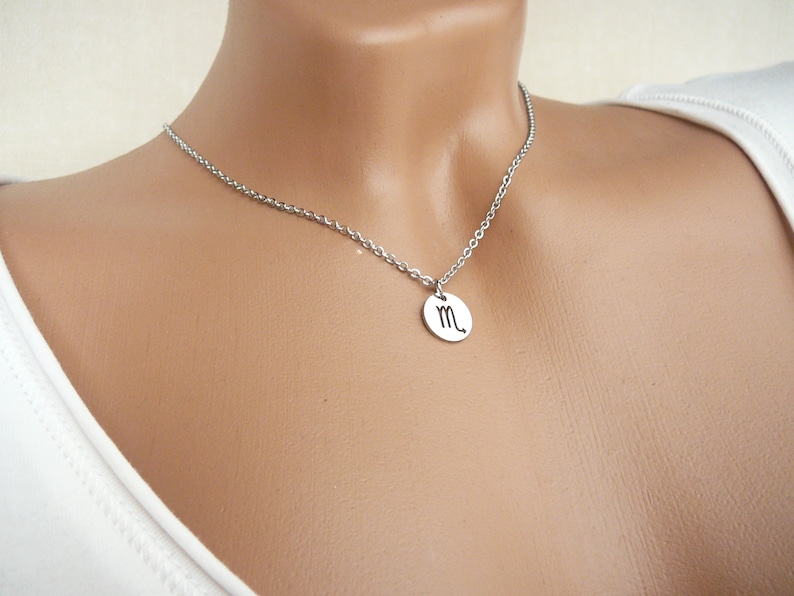 May include: A silver necklace with a round pendant featuring the zodiac symbol for Scorpio.