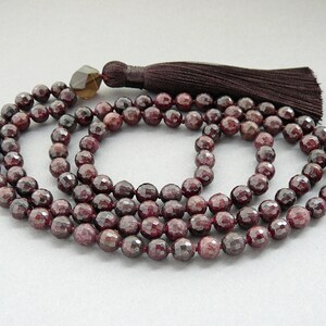 Può includere: Una collana mala di pietre preziose rosse con un fiocco marrone. La collana è composta da 108 perle ed è lunga circa 46 cm.