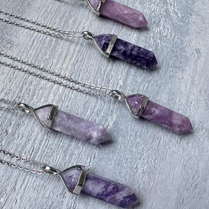 Puede incluir: Cinco collares con colgantes de piedra morada, pulidos y hexagonales, suspendidos de cadenas de metal plateado. Cada colgante est&aacute; asegurado por una tapa de metal plateado, mostrando variados tonos de texturas minerales violetas.