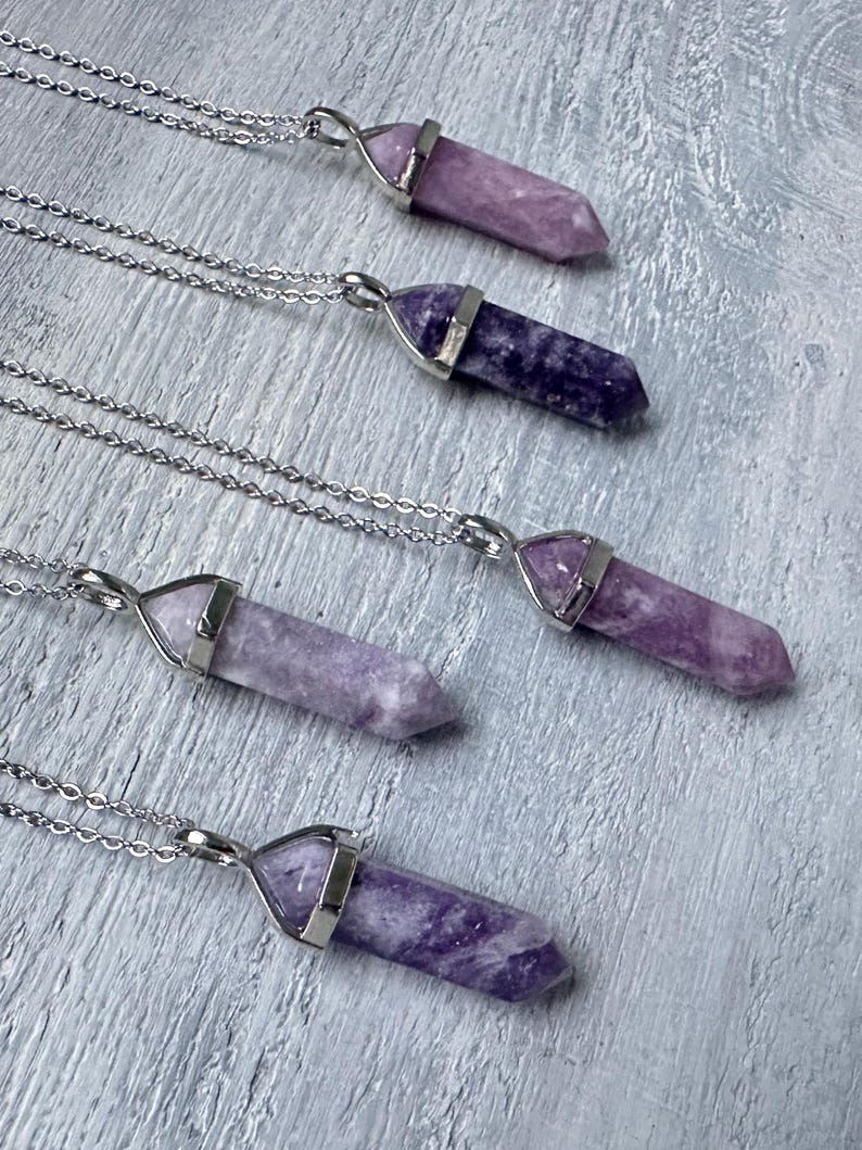 Puede incluir: Cinco collares con colgantes de piedra morada, pulidos y hexagonales, suspendidos de cadenas de metal plateado. Cada colgante est&aacute; asegurado por una tapa de metal plateado, mostrando variados tonos de texturas minerales violetas.