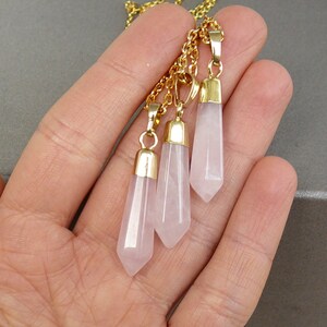 May include: A gold chain necklace with three rose quartz crystal pendants. Each pendant is a pointed, faceted, light pink crystal.