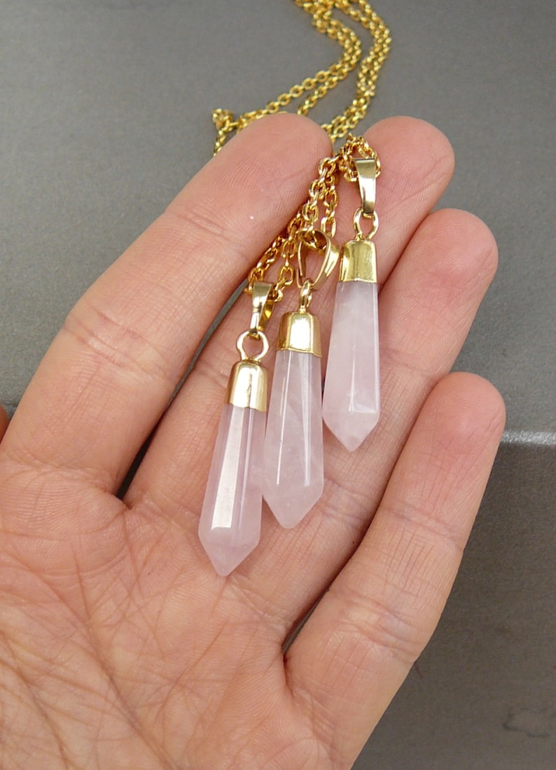 May include: A gold chain necklace with three rose quartz crystal pendants. Each pendant is a pointed, faceted, light pink crystal.