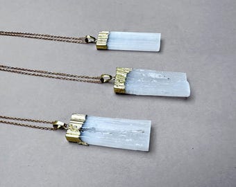 Pode incluir: Três colares de ouro com pingentes de cristal de selenita branca. Os pingentes são retangulares e têm uma moldura dourada. Os colares estão sobrepostos numa superfície cinzenta clara.