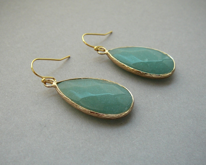 Pu&ograve; includere: Un paio di orecchini a goccia d'oro con una pietra preziosa verde chiaro. Gli orecchini hanno un bordo dorato testurizzato.