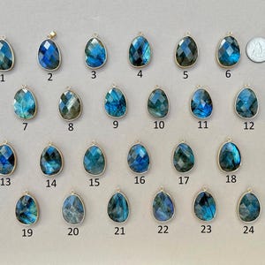 May include: A collection of twenty-four teardrop-shaped labradorite pendants with gold-colored bezels. The pendants display a range of blue and green iridescent colors. A quarter coin is included for size reference. Each pendant is numbered.