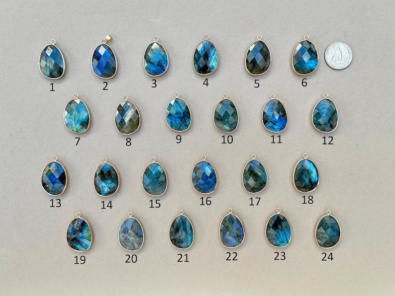 May include: A collection of twenty-four teardrop-shaped labradorite pendants with gold-colored bezels. The pendants display a range of blue and green iridescent colors. A quarter coin is included for size reference. Each pendant is numbered.