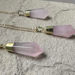 May include: Three gold necklaces with pink crystal pendants. The pendants are shaped like long, pointed teardrops.