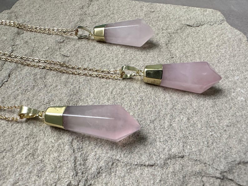 May include: Three gold necklaces with pink crystal pendants. The pendants are shaped like long, pointed teardrops.