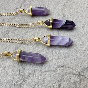 May include: Four necklaces featuring hexagonal amethyst crystal pendants with gold-toned metal caps. Each pendant is suspended from a delicate gold-toned chain. The amethyst stones display natural variations in purple and white banding throughout.