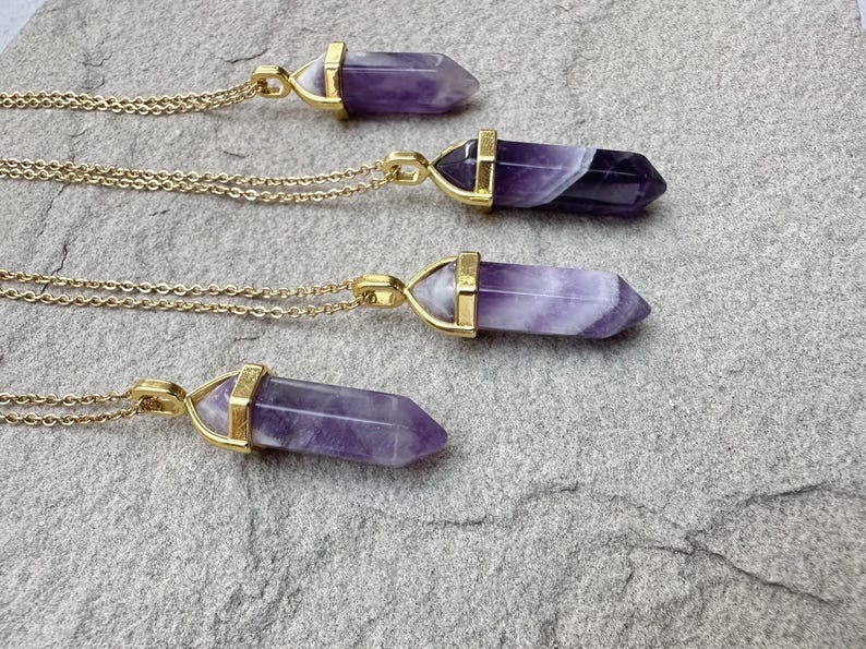 May include: Four necklaces featuring hexagonal amethyst crystal pendants with gold-toned metal caps. Each pendant is suspended from a delicate gold-toned chain. The amethyst stones display natural variations in purple and white banding throughout.