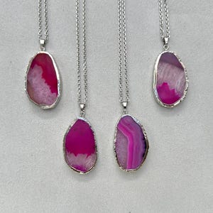 May include: Four silver chain necklaces with agate pendants. The pendants are a mix of pink and white, with a silver trim. The necklaces are displayed on a light grey surface.
