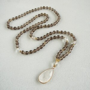 May include: A brown smoky quartz mala necklace with a clear quartz pendant. The necklace is made with 108 beads and has a tassel at the end.