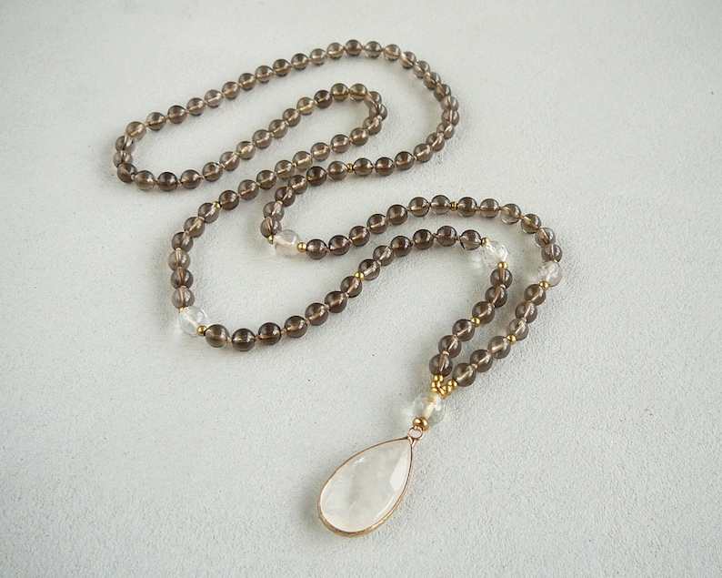 May include: A brown smoky quartz mala necklace with a clear quartz pendant. The necklace is made with 108 beads and has a tassel at the end.
