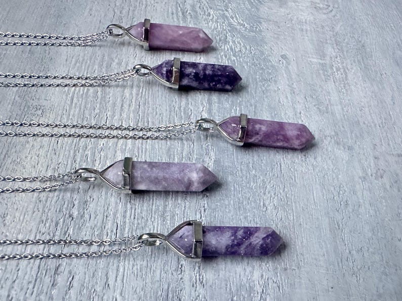 Puede incluir: Cinco collares con colgantes de cristal hexagonal puntiagudos en varios tonos de morado y lavanda. Cada piedra est&aacute; asegurada en una tapa de metal plateado y unida a una delicada cadena plateada, dispuestas en una fila paralela.