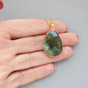 May include: A gold-toned necklace featuring a large, faceted labradorite pendant. The pendant has a teardrop shape and displays shades of green and blue. The necklace is displayed on a light gray background.