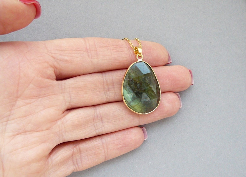 May include: A gold-toned necklace featuring a large, faceted labradorite pendant. The pendant has a teardrop shape and displays shades of green and blue. The necklace is displayed on a light gray background.