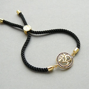 May include: A black adjustable cord bracelet with a gold Om symbol charm. The charm is surrounded by small purple rhinestones.