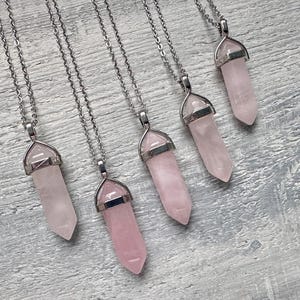 Puede incluir: Cinco collares con colgantes de cristal de cuarzo rosa claro, pulidos y hexagonales. Cada cristal está asegurado en una tapa de metal plateado y cuelga de una cadena plateada a juego. Los colgantes están dispuestos en diagonal.