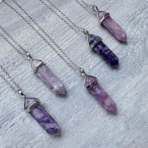 Puede incluir: Cinco collares con colgantes de cristal hexagonal puntiagudo en varios tonos de morado, desde lavanda claro hasta violeta intenso. Cada cristal est&aacute; rematado con una anilla de metal plateado y cuelga de una cadena fina de metal plateado.