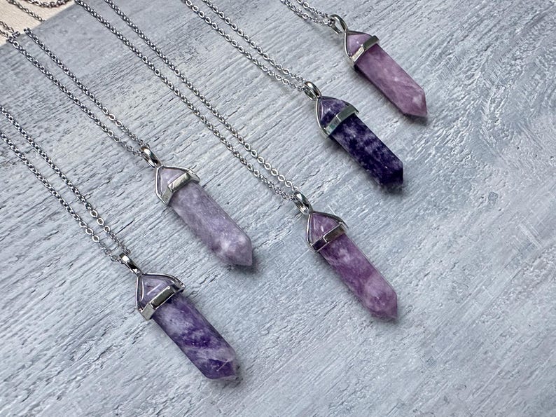 Puede incluir: Cinco collares con colgantes de cristal hexagonal puntiagudo en varios tonos de morado, desde lavanda claro hasta violeta intenso. Cada cristal est&aacute; rematado con una anilla de metal plateado y cuelga de una cadena fina de metal plateado.