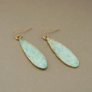 May include: A pair of teardrop-shaped earrings. Each earring features a pale blue stone with a gold-colored metal trim and a gold-colored hook. The earrings are displayed against a neutral background.