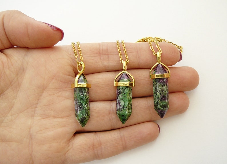 May include: Three gold necklaces with green and purple gemstone pendants. The pendants are shaped like long, pointed crystals with gold caps.