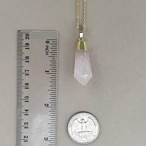 May include: A gold chain necklace with a rose quartz pendant. The pendant is a pointed, faceted crystal that is about 1.5 inches long. The necklace is shown next to a ruler and a US quarter for size reference.