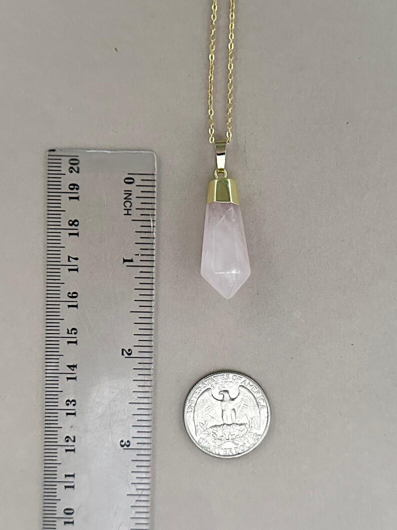 May include: A gold chain necklace with a rose quartz pendant. The pendant is a pointed, faceted crystal that is about 1.5 inches long. The necklace is shown next to a ruler and a US quarter for size reference.