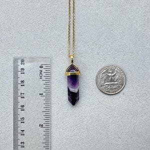 May include: A gold-toned chain necklace featuring a hexagonal, pointed amethyst pendant with a gold-toned metal cap. A ruler shows the pendant is approximately 1.25 inches long, and a US quarter coin is placed next to it for scale.