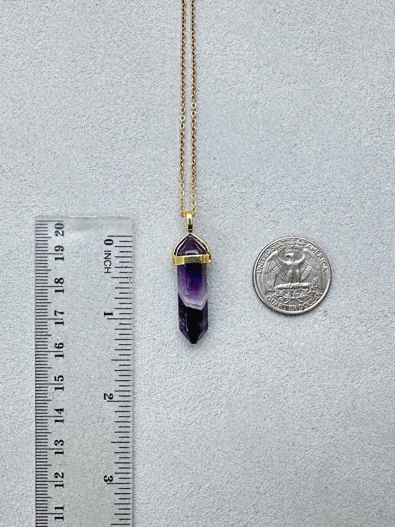 May include: A gold-toned chain necklace featuring a hexagonal, pointed amethyst pendant with a gold-toned metal cap. A ruler shows the pendant is approximately 1.25 inches long, and a US quarter coin is placed next to it for scale.