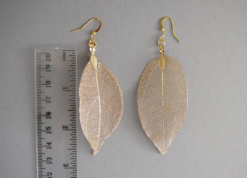 May include: A pair of gold-colored leaf earrings with a delicate, veined design. The earrings are approximately 3 inches long and hang from gold-colored ear wires. The earrings are displayed on a gray background.